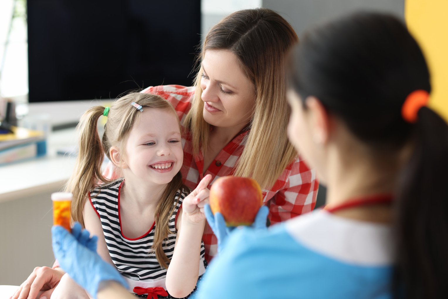 Doctor Offering Little Girl Bottle of Medicine and Red Apple in Clinic